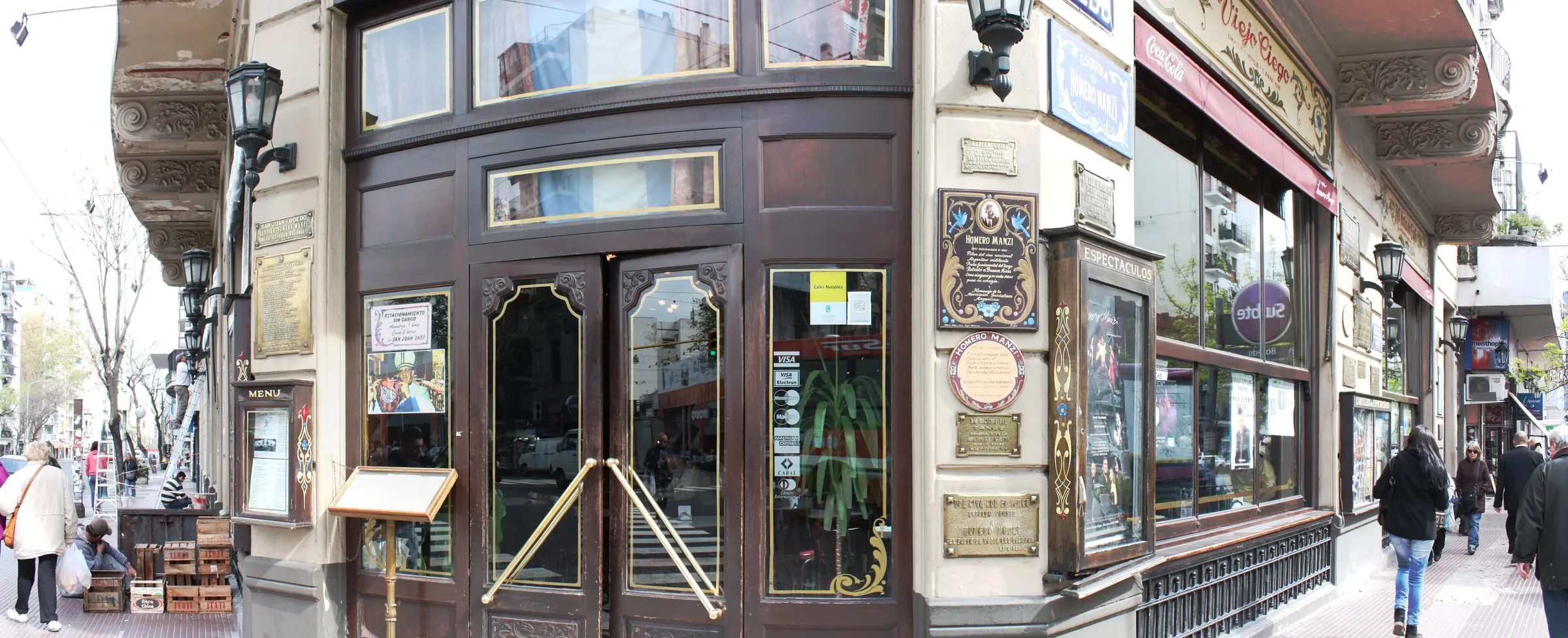 The curved corner entrance of Esquina Homero Manzi at San Juan and Boedo, with dark wooden doors, brass handles, brass plaques and painted signs on the walls either side