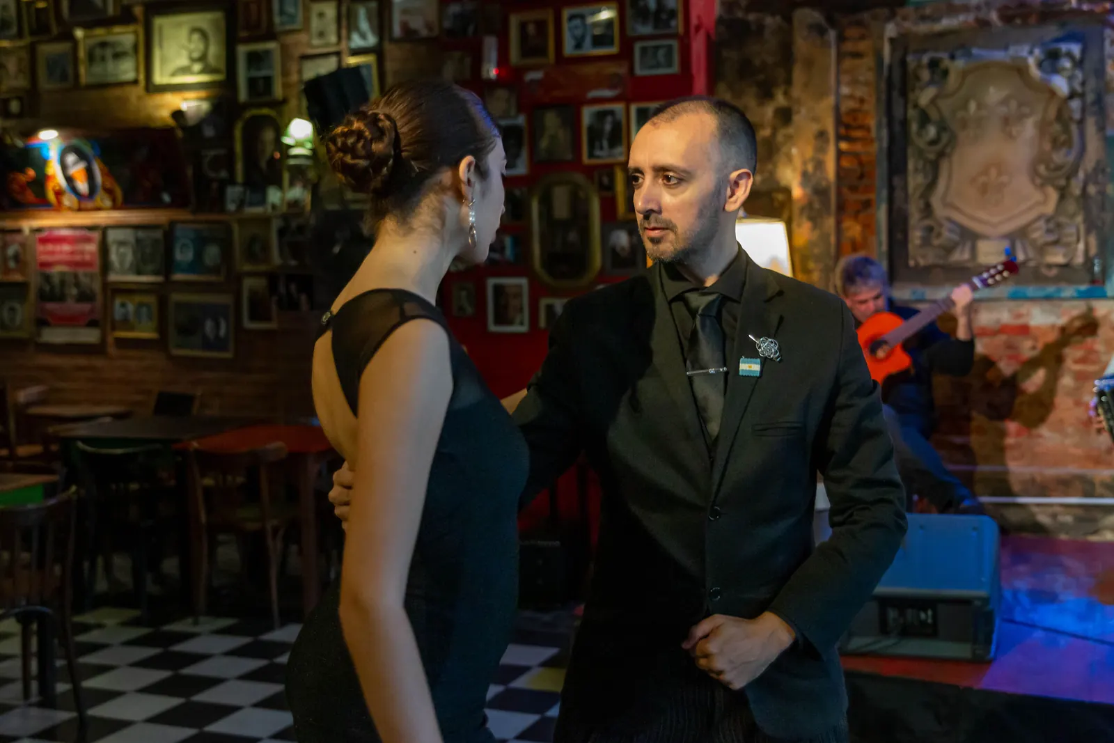 A tango couple dressed in black dancing in a bar in Argentina while a guitarist plays behind them, photographed at a different venue