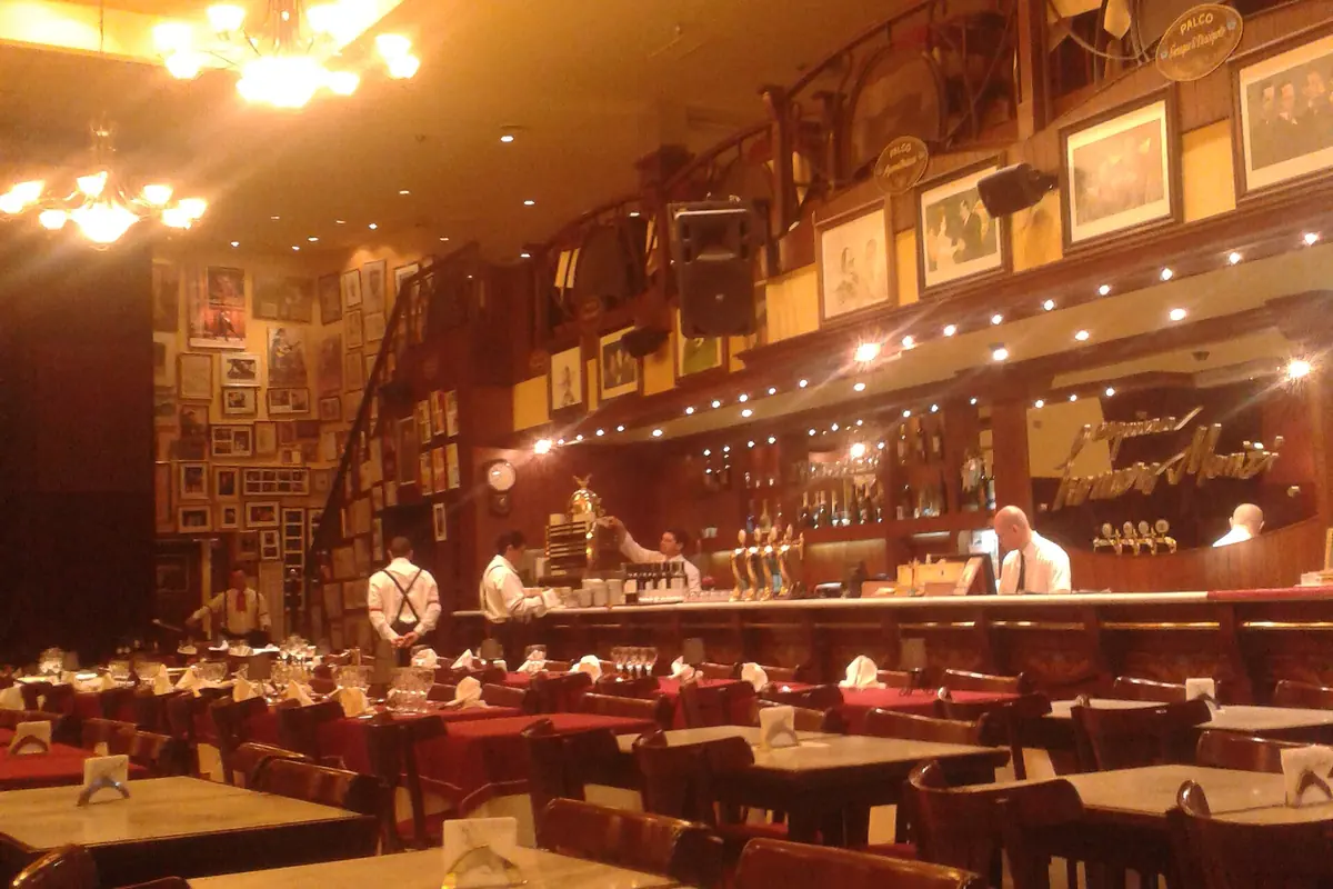The dining hall of Esquina Homero Manzi before service, with marble-topped tables, red cloths, an upper gallery of boxes hung with framed pictures and a long bar