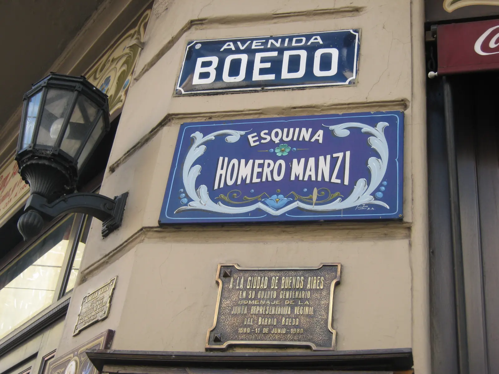 The blue painted corner sign reading Esquina Homero Manzi beneath the Avenida Boedo street sign, with an iron street lamp beside it